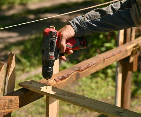 A man drills a hole in a wooden board. Making a fence in the garden.